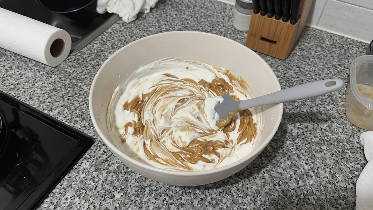 Overhead view of a mixing bowl with Greek yogurt and peanut butter half stirred together, a spatula in the bowl, on a kitchen counter
