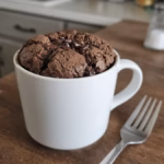 White ceramic mug with a risen chocolate protein mug cake domed slightly above the rim, a few melted mini chocolate chips on top, and a fork beside the mug on a wooden counter